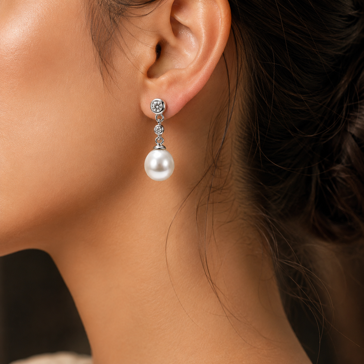 A close-up of a woman's ear wearing a dangling pearl earring with a silver-toned stud and small clear stones.