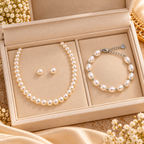 A pearl necklace and matching pearl stud earrings displayed in a beige jewelry box next to a matching pearl bracelet with silver-toned beads and chain, set on a satin fabric background with small white flowers.